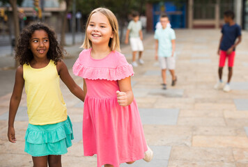 Happy blonde preteen girl walking with her african american girl playmate along city street on summer day .. - obrazy, fototapety, plakaty