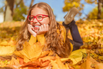 a teenage girl in red glasses lies in yellow autumn foliage and smiles