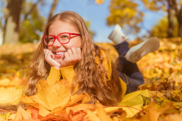 a teenage girl in red glasses lies in yellow autumn foliage and smiles