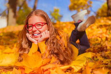 a teenage girl in red glasses lies in yellow autumn foliage and smiles