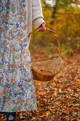 A woman in a dress with a pattern of plants and flowers goes harvesting along a path covered with leaves in fall with a wicker basket with handle