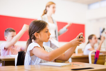 Portrait of schoolgirl using mobile phone while working in classroom during lesson in primary school