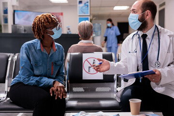 Healthcare professional receiving african american patient in hospital room. Doctor attending sick woman in clinic waiting area. Medical staff, people wearing masks at sanatorium reception. - obrazy, fototapety, plakaty