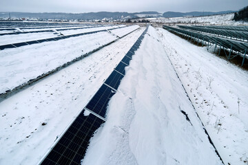 Aerial view of snow covered sustainable electric power plant with rows of solar photovoltaic panels for producing clean electrical energy. Low effectivity of renewable electricity in northern winter