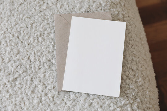 Neutral stationery still life scene. Blank greeting card, invitation mockup and craft envelope on white boucle sofa upholstery. Teddy textured fabric. Blurred oak wooden parquet floor. Top view.