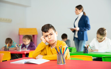 Portrait of upset boy in schoolroom on background with pupils studying with teacher