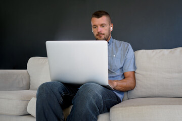 Man working at home using laptop with wireless earphones, sitting on couch.