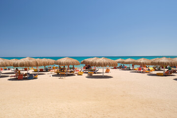 Straw shade umbrellas on sea tropical beach with resting sunbeds against blue vibrant sky in summer