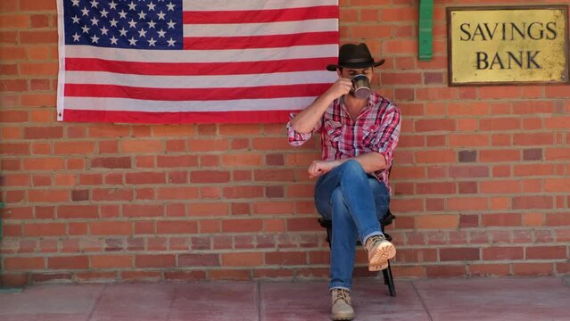 male cowboy sitting drinking whiskey from a mug in a saloon . a cowboy in a plaid shirt in the Populated western town sits at a tavern and counts money. Street of wild west town with bank building