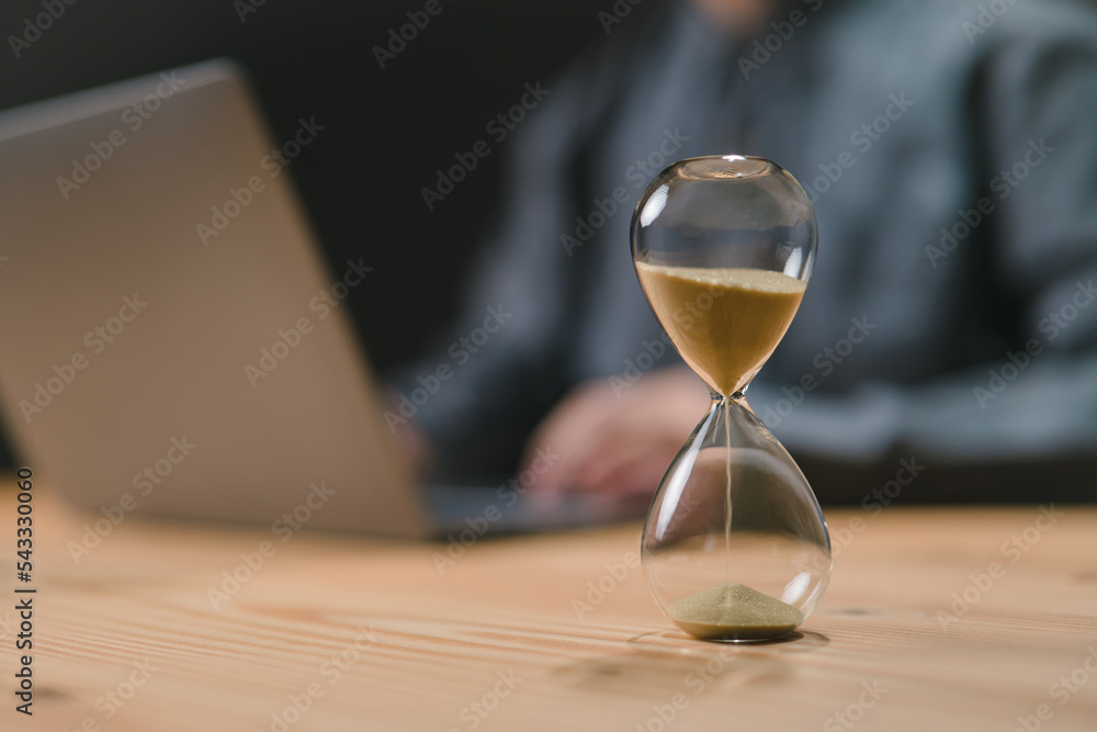Poster hourglass stand on a wood desk in the man home office, measuring time while he is busy using a compu