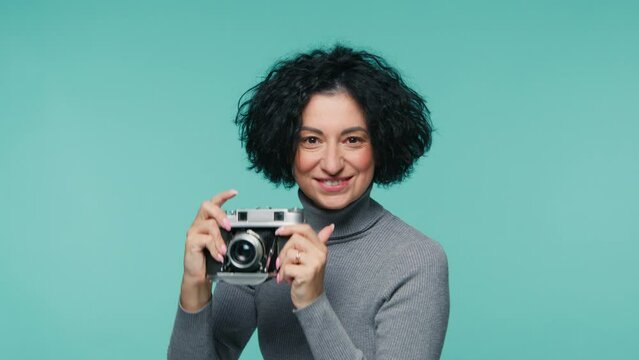 Proud mature photographer female 40s holding analog camera and taking pictures on pastel blue background. Smiling adult woman with short haircut looking at camera and taking picture on film camera
