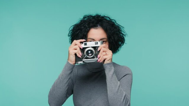 Smiling adult woman with short haircut looking at camera and taking picture on film camera. Proud mature photographer female 40s holding analog camera and taking pictures on pastel blue background