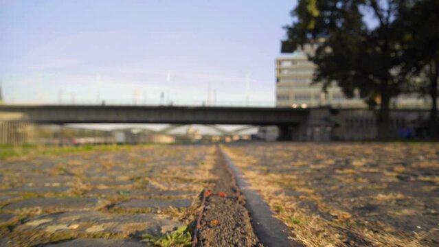 Low dolly shot of old railroad tracks in cobble stones with grass and weeds growing up around the edges. Tight focus with a bridge and people walking in the background