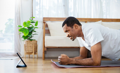 Fototapeta premium Portrait of senior mature African black man warm up exercise workout yoga plank position at home living room. Yoga for beginner. Guy watching online tutorials on tablet, training, healthy quarantine