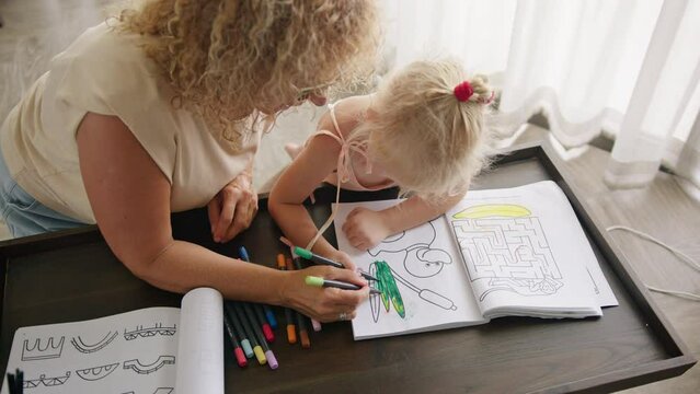 Curly mother plays with her daughter and teaches her to draw with pencils in a coloring book, sitting on the floor near the table. The concept of home education and togetherness with the family.
