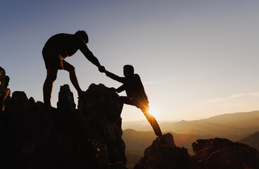 Fototapeta Silhouette of  two people climbing on mountain cliff and one of them giving helping hand. People helping and, team work concept. obraz