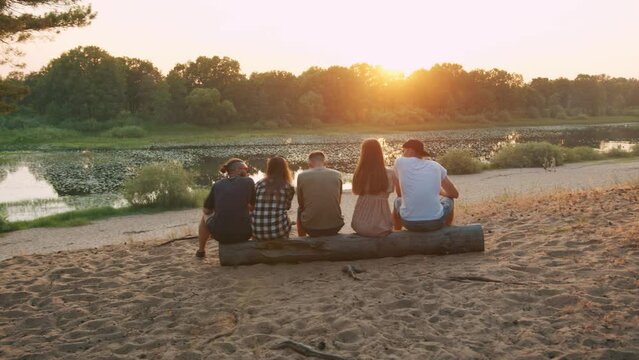 A group of guys and young girls are sitting on a log on the river bank and talking. View from the back. Slow motion