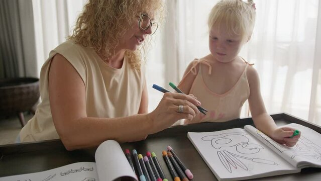 Grandmother plays with her granddaughter and teaches her to draw with bright pencils in a coloring book, sitting in the living room. The concept of home education and togetherness with the family.
