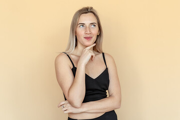 Fototapeta premium Dreamy pretty young woman with freckles touch chin thought choose decide solve problems dilemmas, wear fashionable top, stands over beige background. Thinking positive concept
