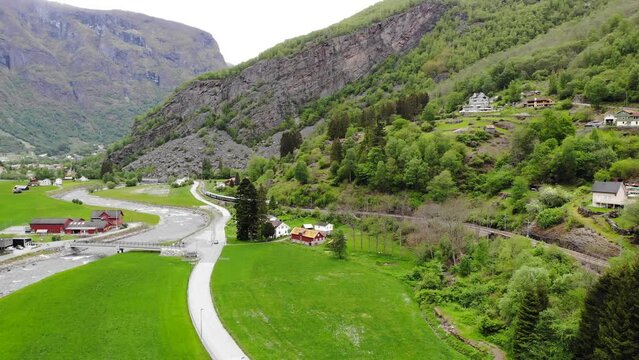 Aerial: Flåm Train Going Uphill Through A Valley Among Green Meadows And A Road With Light Traffic