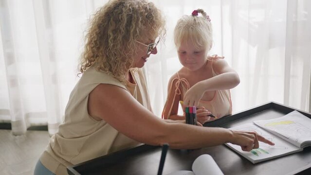 A smiling curly-haired grandmother teaches her granddaughter different colors and chooses a pencil to draw with. The concept of home education and togetherness with the family.