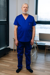Full length vertical portrait of tired mature adult male doctor in medical uniform standing in hospital office on background of window, looking at camera. Gray-haired practitioner posing at workplace.
