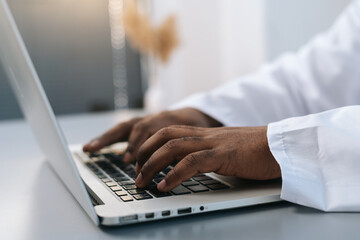 Close-up side view hands of unrecognizable African-American male doctor wearing white coat working typing on laptop computer, sitting at desk in medical office room at hospital near window.