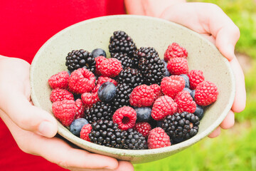 Boy holding green bowl with freshly picked garden berries with his hands - raspberry, blackberry, blueberry. Healthy eating and dieting concept. Organic superfood snack. Summer berries background