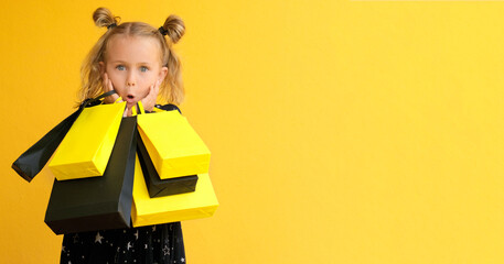 Surprise child girl in dress hold black package bags with purchases isolated on yellow background studio. Shopping online discount sale. Mock up,copy space, banner. Black friday