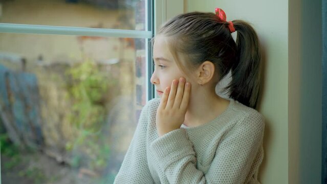 Scared sad girl refugee sits on windowsill and looks out window at curfew because russian armored invasion into Ukraine. Save lifes, no war, world peace, stop conflict, born love. Childhood in