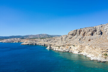 Fototapeta na wymiar Aerial view of a peaceful rocky coastline and beautiful turquoise waters with mountain range. Mediterranean Sea, Rhodes Island, Greece.