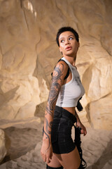 armed and tattooed archaeologist in sexy outfit looking away while walking in cave.