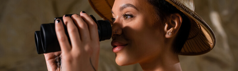 young archaeologist in safari hat looking through binoculars in desert, banner.
