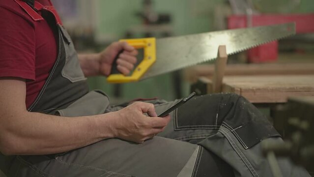 a man saws a board with a wood saw and looks into his smartphone. slow motion video