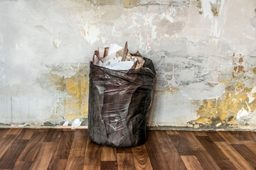 A black trash bag filled with old torn wallpaper stands against a plastered wall in an apartment....