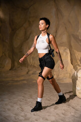 full length of sexy and tattooed archaeologist with holster and gun walking on sand in cave.