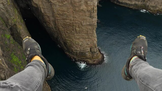 4K Beautiful View Of The Sorvagsvatn Lake Above The Ocean And Men Legs Hanging Down From The Cliff In Vagar, Faroe Islands. Perpendicular Rock Wall. Legs Man Sitting Edge Cliff.