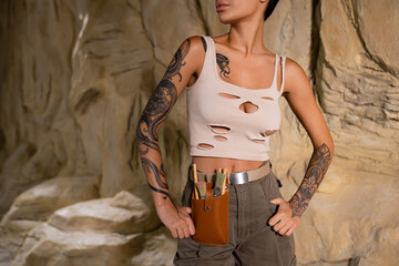 partial view of tattooed archaeologist in tank top and waist bag with brushes standing with hands on hips near rock. - Powered by Adobe