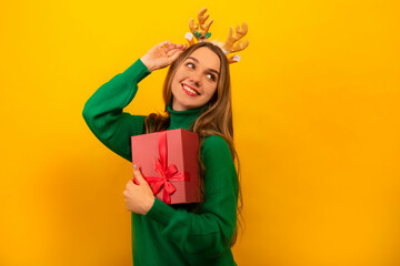 Happy young woman wearing Christmas toy reindeer antlers and green sweater, holding pink present or...