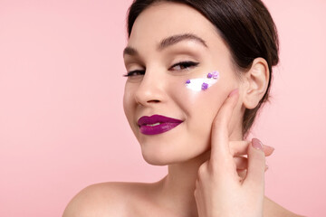 Smiling young woman with  smooth skin, cream and flowers on cheek posing on pink studio background. Stunning female touch  face. Natural woman beauty, skincare cosmetics ad.