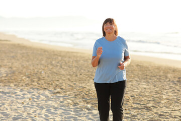 Smiling middle aged woman running on the beach on sunrise. 40s or 50s attractive mature lady in sports clothes doing jogging workout enjoying fitness and healthy lifestyle at beautiful sea landscape.