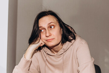 A thoughtful girl in a pink hoodie sits on a chair and looks into the window. Front view, close up