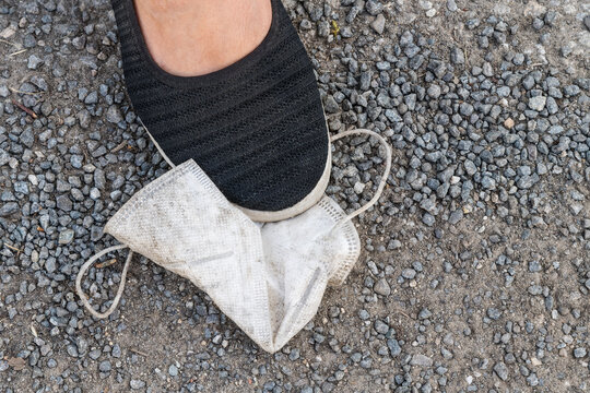 Female foot in black shoe and dirty respirator fallen on street gravel sidewalk background. Human leg on crushed stone road and old white face mask. End of freedom restriction at coronavirus pandemic.