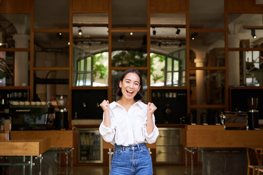 Happy Enthusiastic Asian Girl, Business Owner Triumphing, Opening Her Own Restaurant, Standing In Front Of Empty Cafe