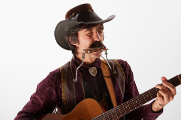Portrait of man with moustaches in country style clothes playing guitar and harmonica, performing isolated over white background