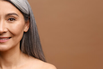Close-up portrait of half face of cheerful middle-aged Asian 50s woman with silver gray long hair, attractive lady looks at camera and laughing. Mature model with naked shoulders isolated on brown