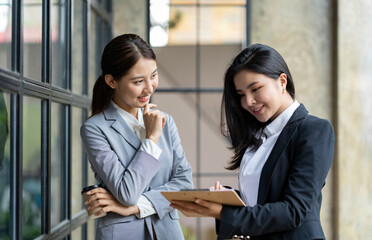 Obraz premium Two pretty young Asian businesswomen standing to discuss working on investment projects and planning strategies together at the office. brainstorming concept.