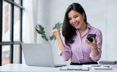 Asian businesswoman raising her hand with a happy expression and looking at her phone The...