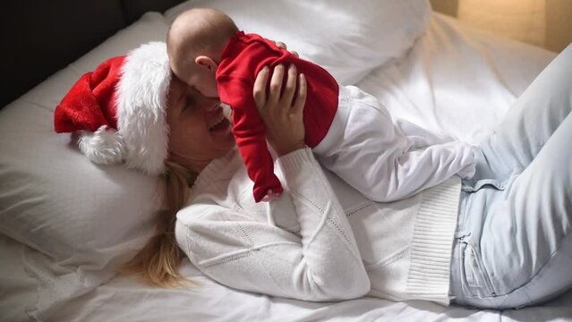 Happy mother lies on bed in red Santa hat and white T-shirt and holds on hands baby in red T-shirt . Newborn - happiness for family. New year and Christmas. elf and fairy. Side view.
