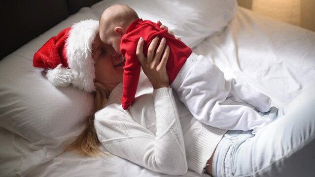 Happy mother lies on bed in red Santa hat and white T-shirt and holds on hands baby in red T-shirt . Newborn - happiness for family. New year and Christmas. elf and fairy. Side view.